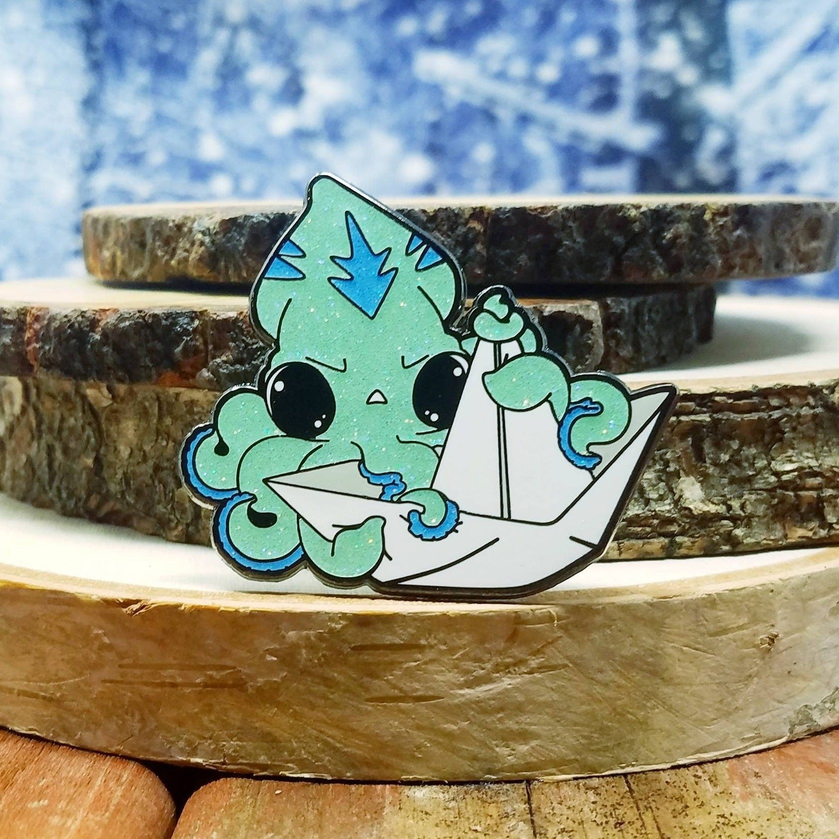 A photograph of a pile of birch rounds with a winter background. In front of the birch rounds is an enamel pin of a light blue squid grabbing onto a paper boat.
