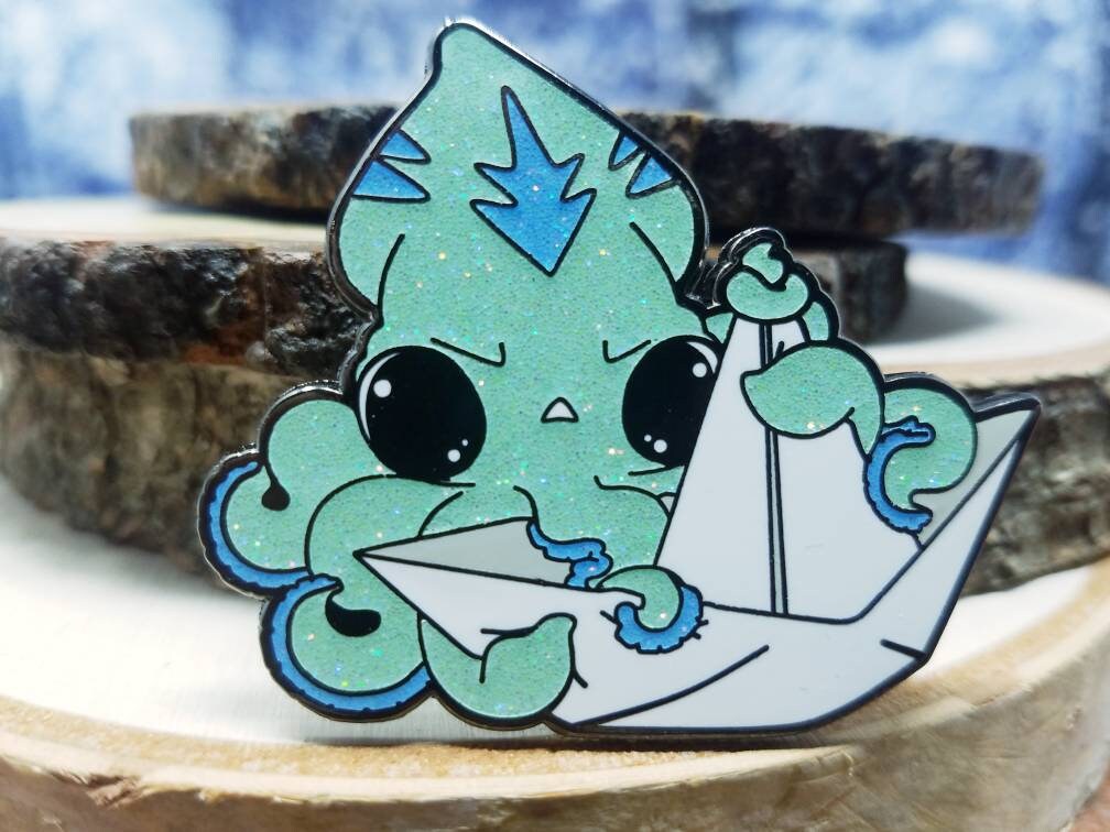 An enamel pin of a light blue squid grabbing onto a paper boat.