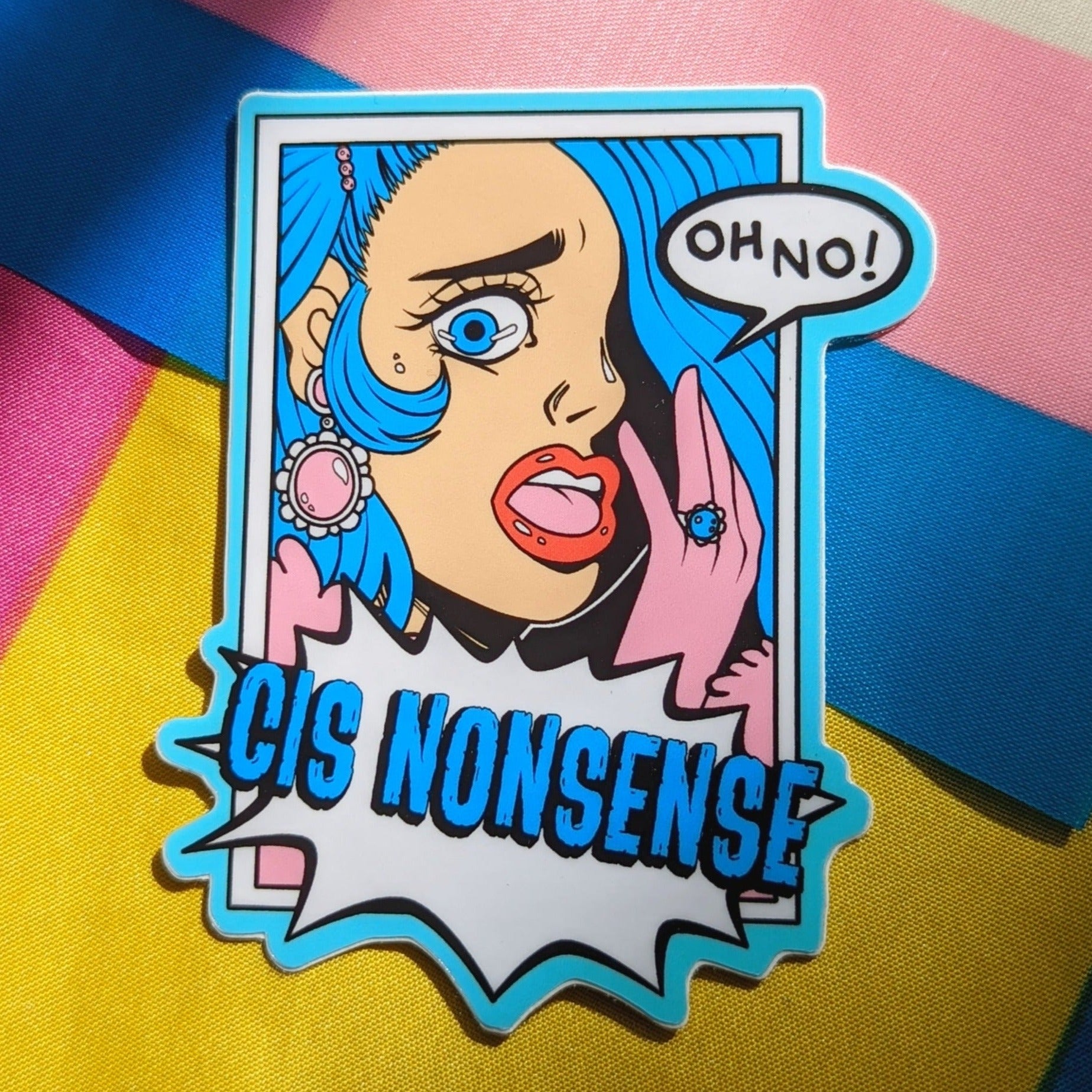 A photograph of a background with the transgender and pansexual pride flags. On top of the flags is a vinyl sticker of a blue haired person with pink gloves gasping and saying “OH NO!” under them is an action bubble that says “CIS NONSENSE.”