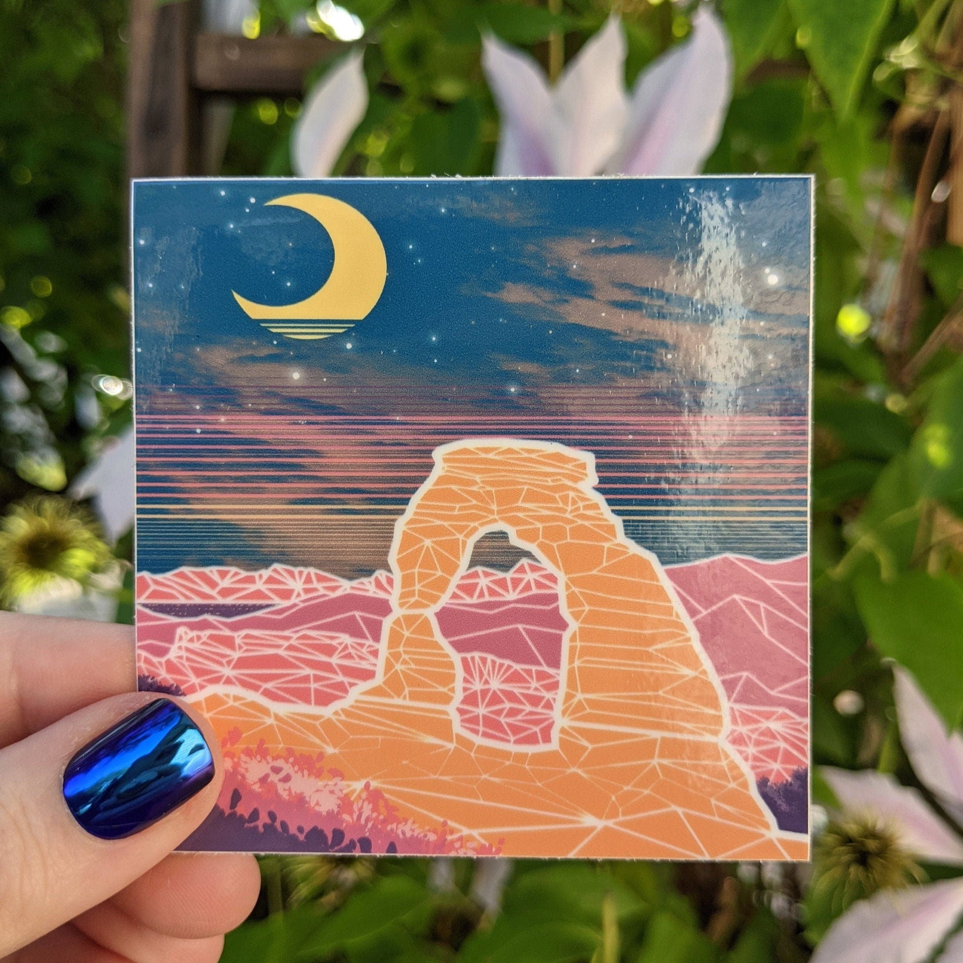 A photograph of a vinyl sticker being held in front of a clematis plant. The vinyl sticker is of delicate arch at Arches national park at night with a vaporwave overlay.