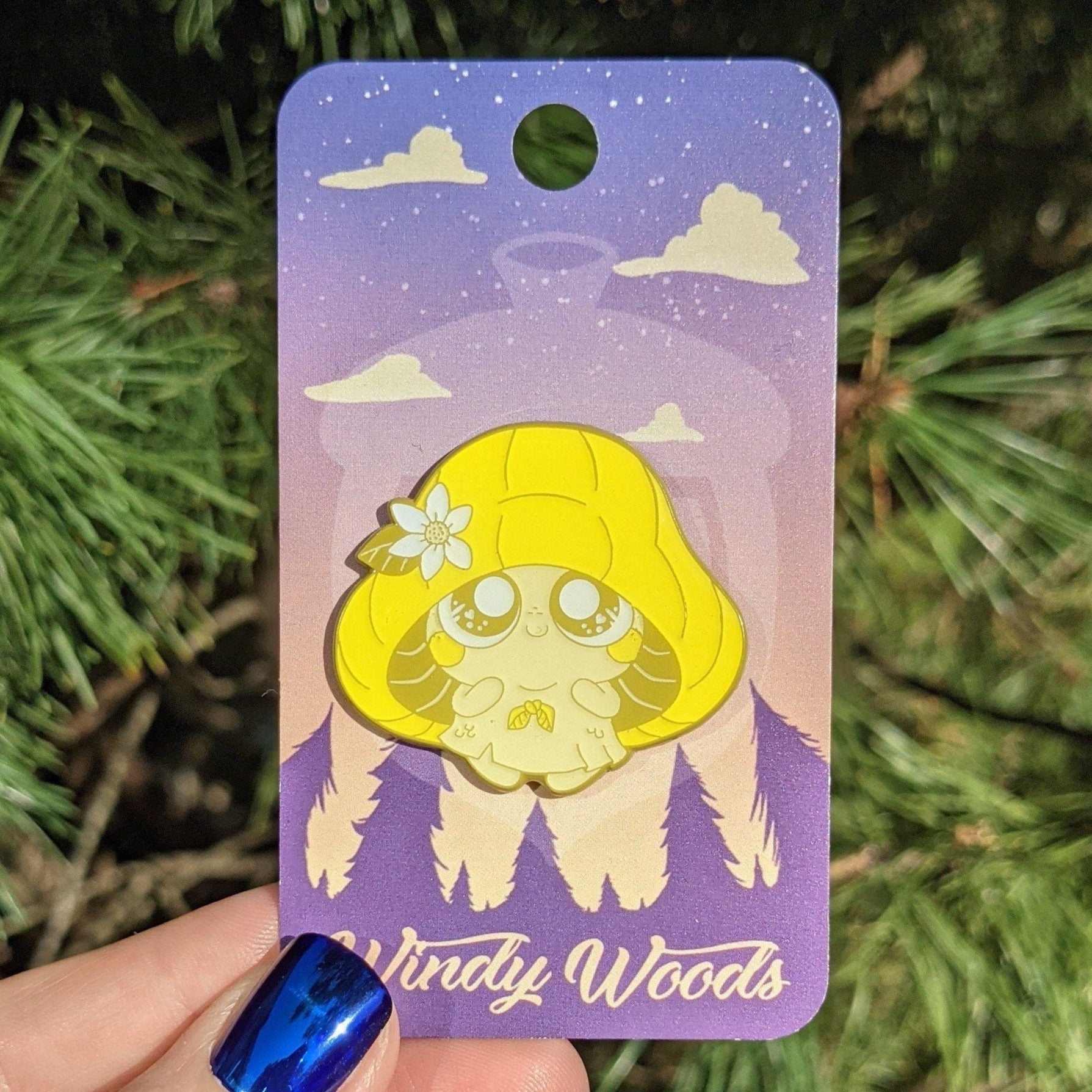 A photograph of an enamel pin being held in front of a pine tree. The enamel pin is on a purple Windy Woods backing card. The pin is a yellow mushroom sprite called Mushkin.
