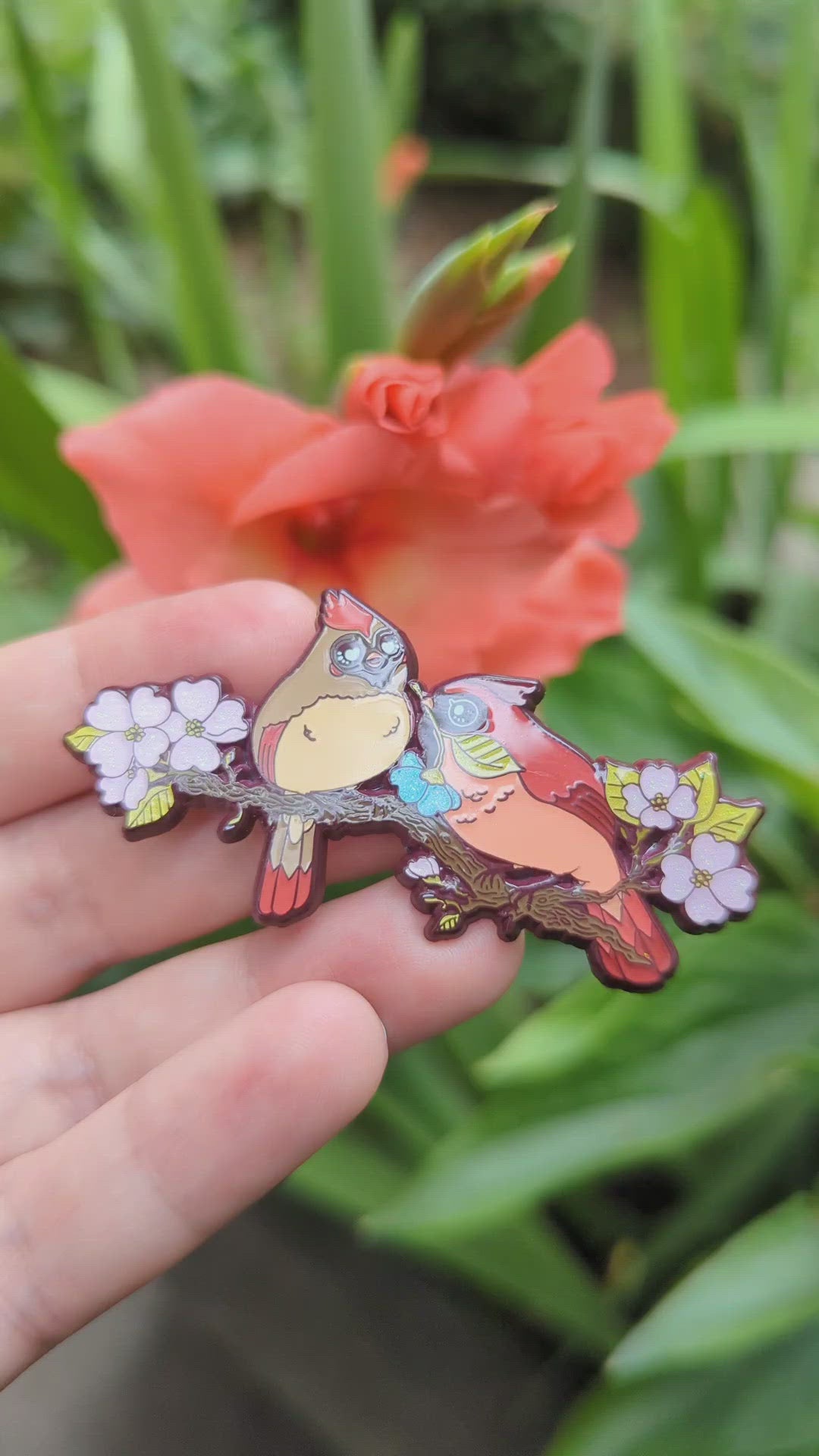 A photograph of a person holding an enamel pin in front of an orange flower. The enamel pin shows two cardinals on a dogwood tree surrounded by pink flowers. One of the cardinals is handing the other a special blue flower.